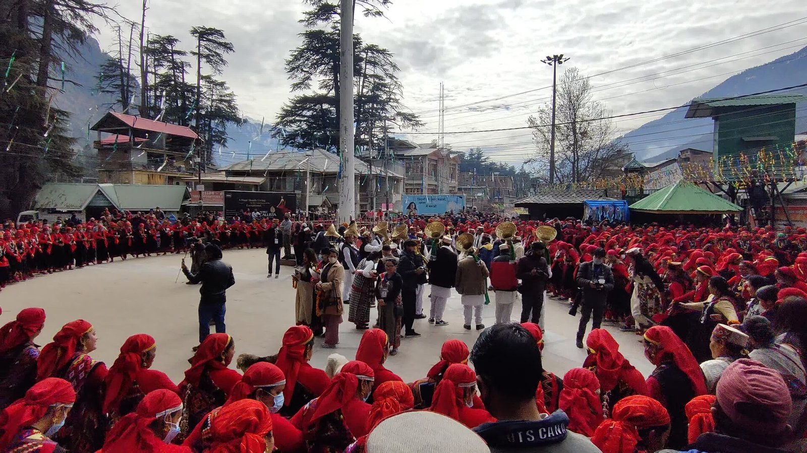 Massive circle of women in red dhattu caps and traditional Kullu dress filling a snow-fringed Manali ground, brass-instrument musicians in the centre, deodars on the hill behind