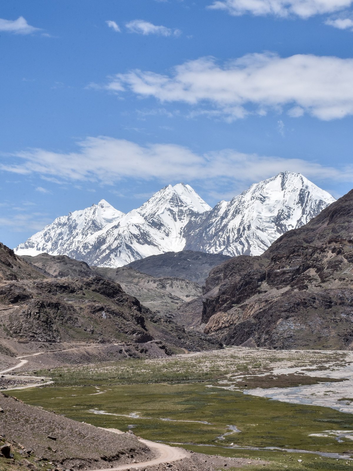 Narrow dirt track climbing through a green meadow toward Kunzum La pass with snow-covered Himalayan peaks rising behind, single tyre-groove visible in the gravel surface