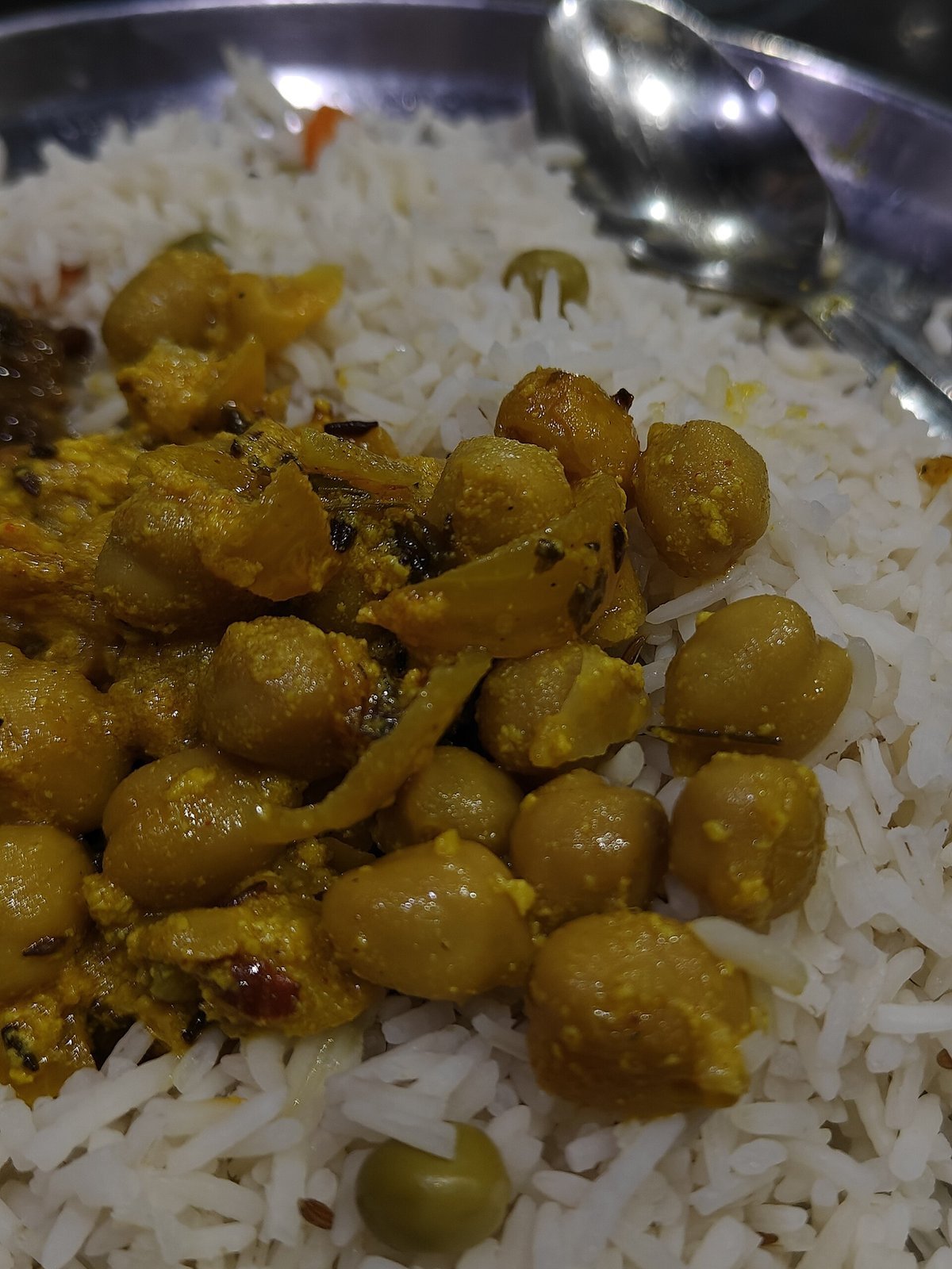 Chana madra served over rice on a steel thali — white chickpeas in yellow yoghurt-spice gravy, no onion, no garlic, with green peas visible, a spoon to one side, the signature dish of Kangri Dham