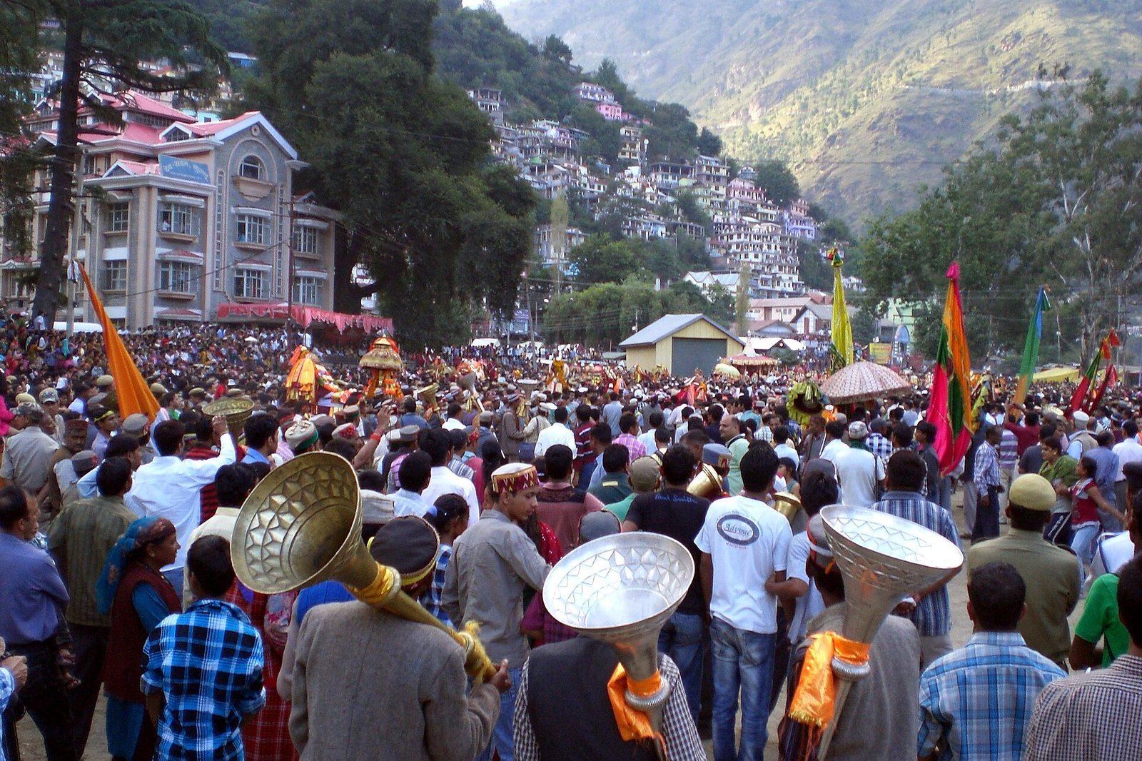 Wide-angle Kullu Dussehra crowd at Dhalpur Maidan with deity palanquins moving above heads, brass instruments in the foreground, mountain town of Kullu rising up the slope behind