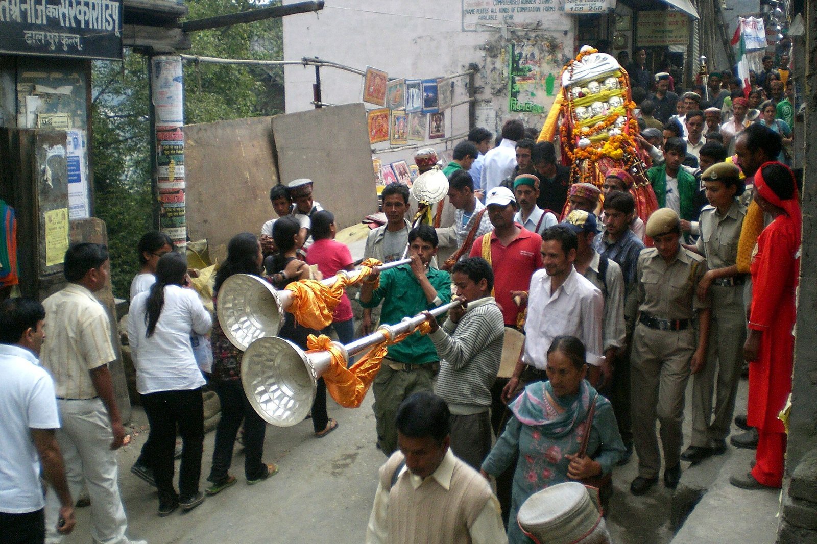 A Kullu Dussehra street procession with two musicians blowing long silver karnal horns wrapped in saffron and orange cloth, a drummer behind, a marigold-draped palanquin carried by the crowd, police providing crowd control